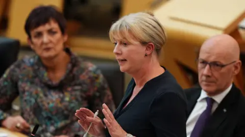 PA Media Shona Robison speaking in the Scottish Parliament, watched by first minister John Swinney.  